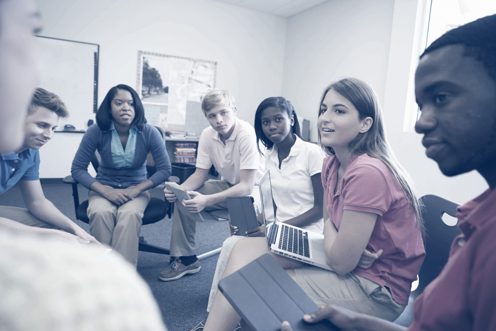 teen students in collared shirts seated in chairs arranged in a classroom discussion circle with laptops