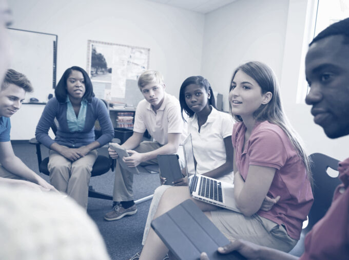 teen students in collared shirts seated in chairs arranged in a classroom discussion circle with laptops